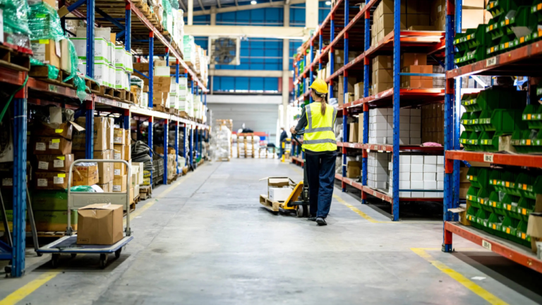 Warehouse worker in safety gear moving boxes on a pallet jack through organized aisles filled with packaged goods and inventory on metal shelving.