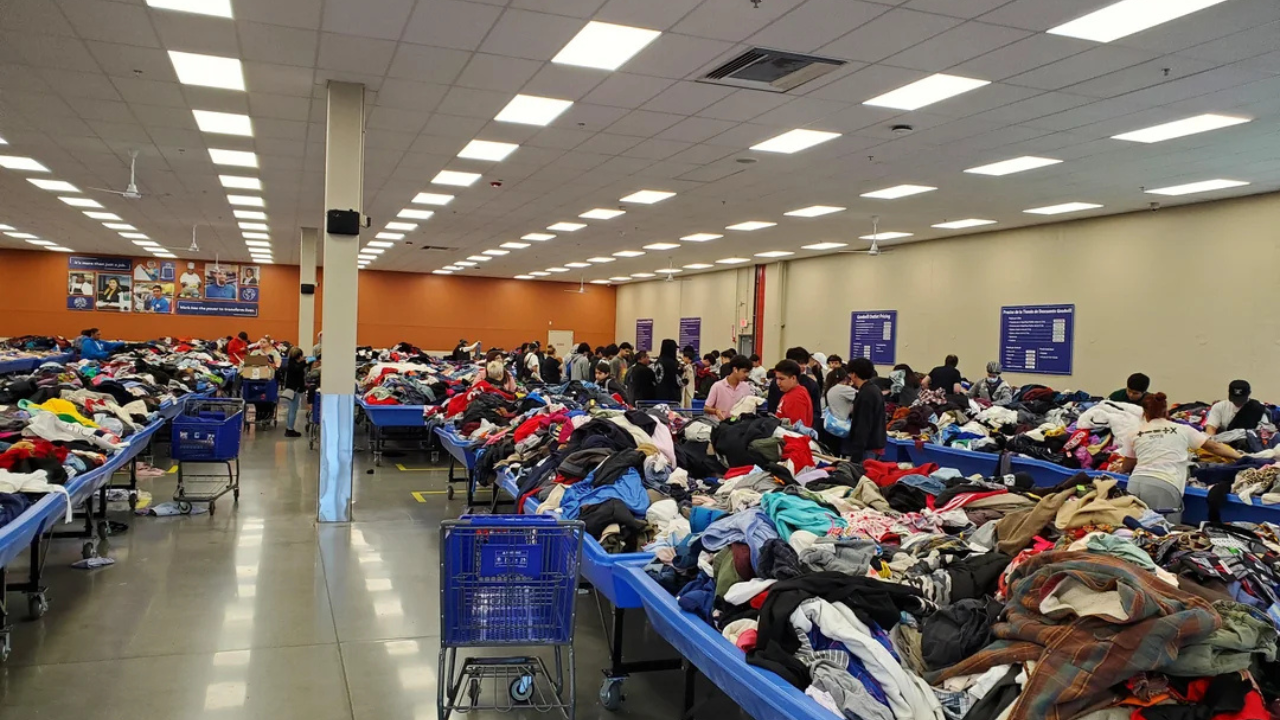 Shoppers browsing through large blue bins overflowing with assorted clothing at a busy bin store with high ceilings and bright overhead lighting