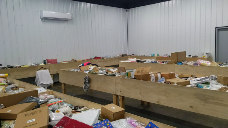 Large wooden bins inside a bin store filled with assorted discounted merchandise such as boxes, clothing, and electronics, set against a plain white metal wall.