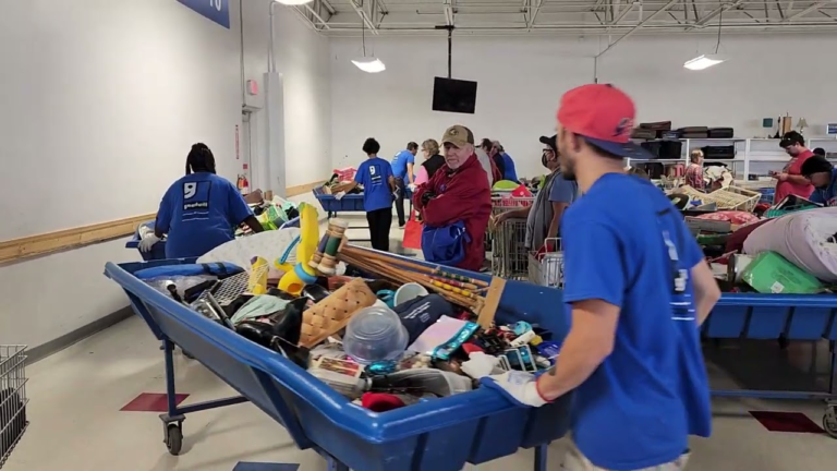 Goodwill employees and shoppers gather around large blue bins filled with assorted secondhand goods, including toys, household items, and electronics, inside a bustling outlet store.