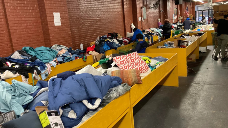 Shoppers browse through large yellow bins filled with assorted clothing, pillows, and miscellaneous household items inside a brick-walled liquidation or bin store.