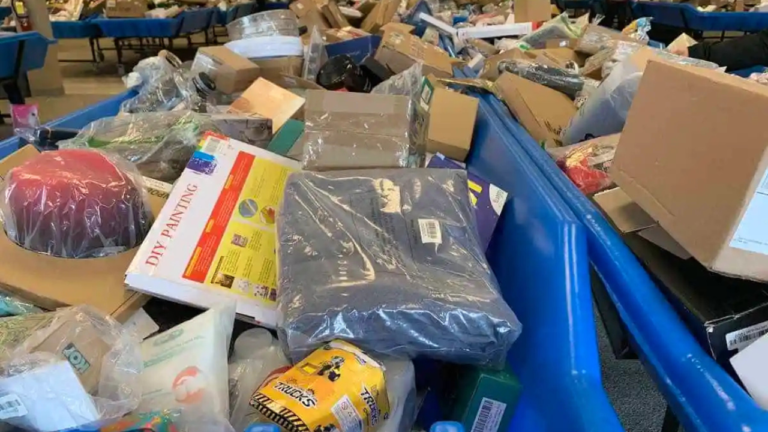 Close-up view of blue bins overflowing with assorted discounted items in a bin store, including boxed goods, toys, a DIY painting kit, clothing in plastic packaging, and miscellaneous household products.