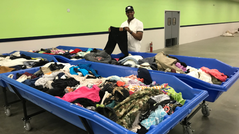 Man standing in a bin store holding up a pair of pants, surrounded by large blue rolling bins filled with assorted secondhand clothing.