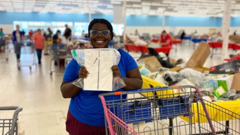 A smiling shopper in a blue shirt holds up a packaged tablet, likely an iPad, inside a busy bin store filled with yellow bins and shopping carts.