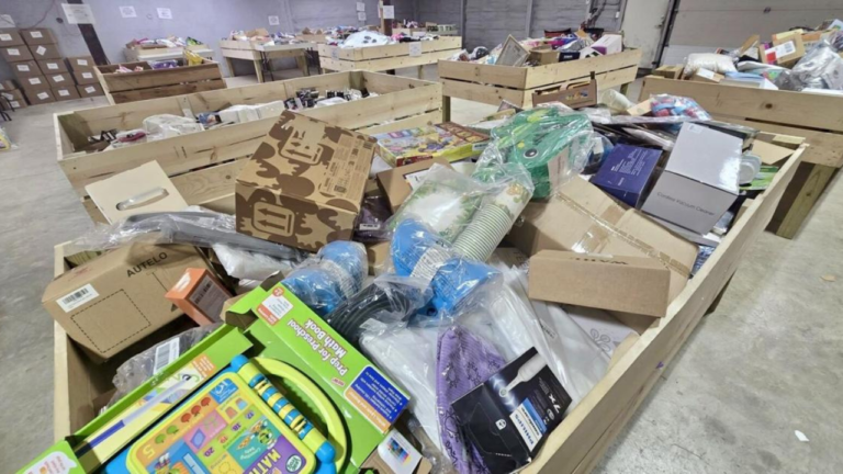 Wooden bins filled with assorted retail items in a warehouse-style bin store, including boxed electronics, toys, and household goods.