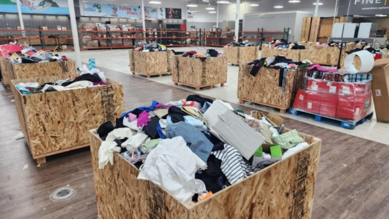 Large wooden bins filled with assorted clothing and goods are spread across a retail warehouse floor, creating a bargain-hunting atmosphere under bright lighting with signs of markdowns and falling prices in the background.