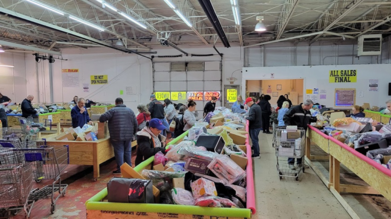 Shoppers browse through large bins filled with assorted merchandise in a warehouse-style discount store, with signs reading “All Sales Final” and “Do Not Open Packages” displayed on the walls.