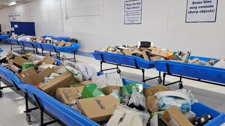 A bin store interior with large blue bins filled with assorted boxed items, electronics, and household goods. Warning signs on the wall advise customers not to open boxes and to beware of sharp objects.
