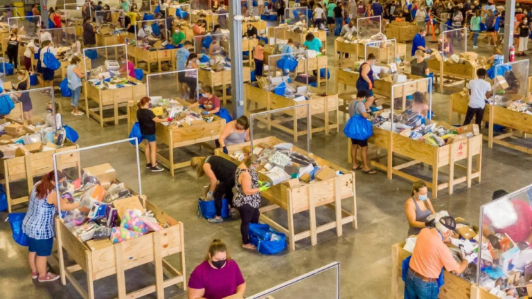 A crowded warehouse-style bin store filled with shoppers digging through wooden bins of assorted merchandise. Many people are carrying large blue bags and wearing face masks.