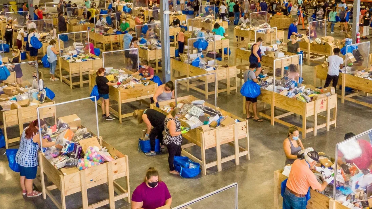 A crowded warehouse-style bin store filled with shoppers digging through wooden bins of assorted merchandise. Many people are carrying large blue bags and wearing face masks.