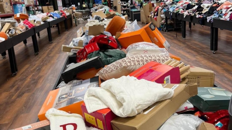 A bin store table overflowing with assorted home goods, clothing, and boxed items, offering a chaotic but treasure-filled bargain shopping experience on a wood-paneled floor.