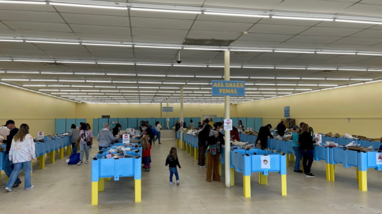 Shoppers browsing through blue and yellow bins filled with assorted merchandise inside a brightly lit bin store with "All Sales Final" signs overhead.