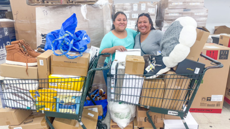 Two smiling women standing behind shopping carts filled with boxes, shoes, household items, and various merchandise at a bin store, with stacked pallets in the background.