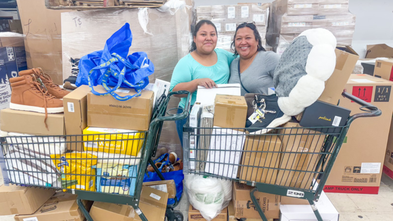 Two smiling women standing behind shopping carts filled with boxes, shoes, household items, and various merchandise at a bin store, with stacked pallets in the background.