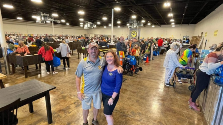 Smiling couple poses at the front of a bustling bin store, surrounded by shoppers digging through large bins filled with discounted items under bright lights and an energetic atmosphere.