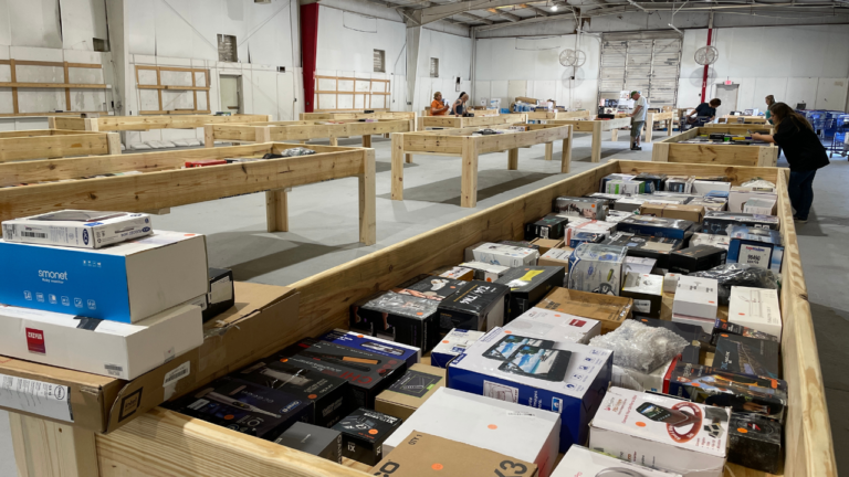 Shoppers browsing through wooden bins filled with various boxed electronics and gadgets inside a spacious bin store with concrete floors and high ceilings.