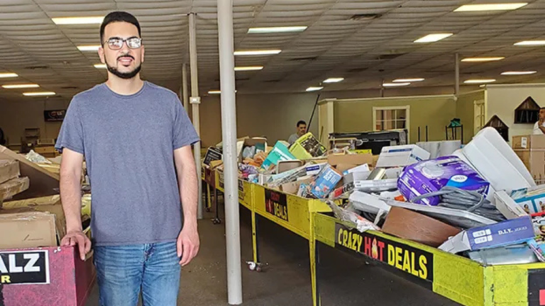 A man standing inside a bin store filled with various discounted merchandise, including boxes of assorted products on tables labeled 'Crazy Hot Deals'.