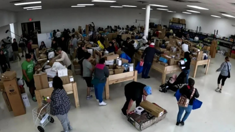 Shoppers search through wooden bins overflowing with discounted goods in a busy bin store, surrounded by boxes and bargain hunters under bright fluorescent lighting.