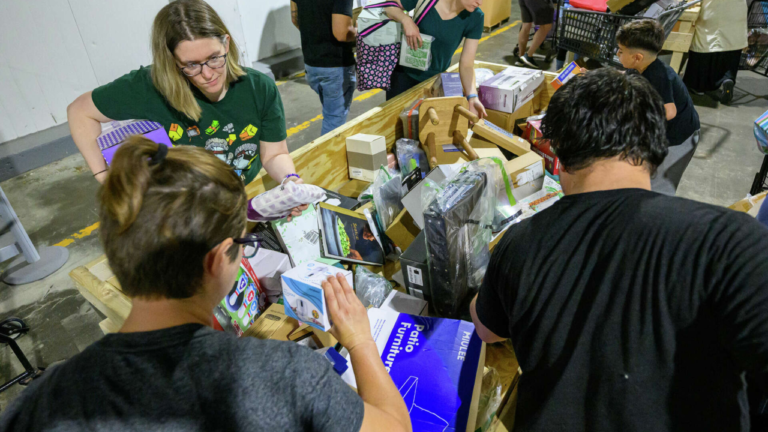 ChatGPT said: Shoppers browsing through assorted discounted merchandise in a wooden bin at a busy bin store, examining items like electronics, books, and home goods.