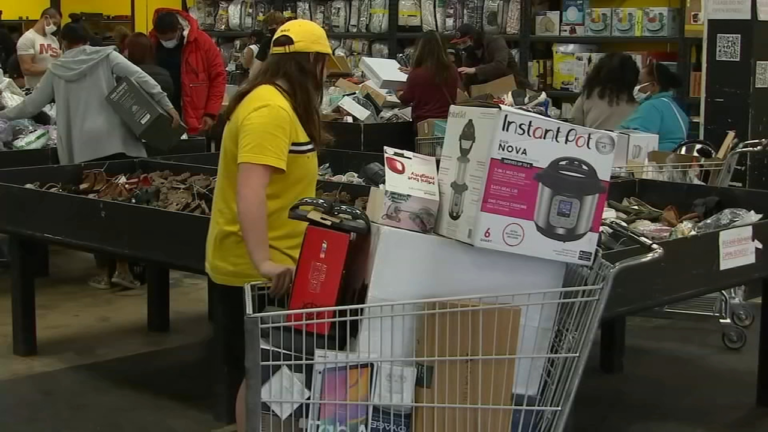 "Shoppers browsing through merchandise bins at a discount store, with one person pushing a shopping cart filled with boxed items including an Instant Pot and electronics."