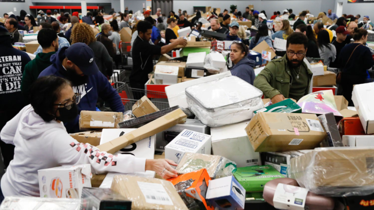 Shoppers searching through piles of discounted merchandise and sealed boxes at a crowded bin store, filled with people looking for deals on various products.