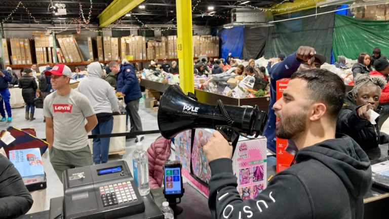 "Man announcing through a megaphone inside a busy bin store, with shoppers browsing through bins filled with various discounted items and boxes in a warehouse-like setting."