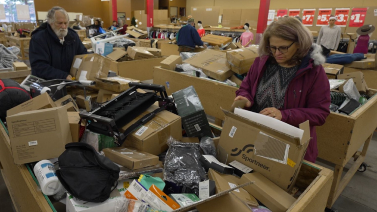 Shoppers browsing through large wooden bins filled with assorted boxes, packaged items, and miscellaneous products inside a bin store, with price signs visible in the background.