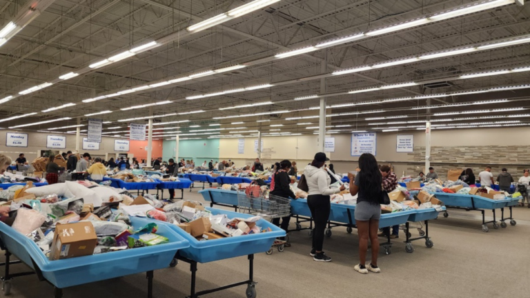 "Shoppers browsing through large blue bins filled with assorted merchandise in a spacious, well-lit bin store with multiple aisles and pricing signs hanging from the ceiling."