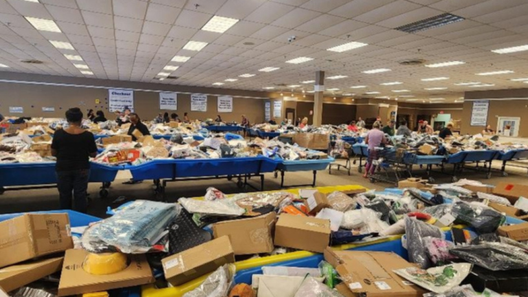 "Shoppers browsing through large blue bins filled with assorted merchandise, boxes, and products inside a spacious bin store with fluorescent lighting and signage indicating checkout and pricing."