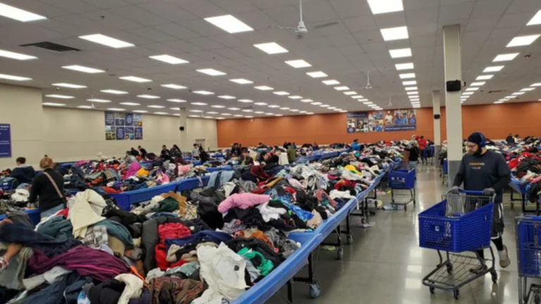 Shoppers browse through large blue bins overflowing with secondhand clothing inside a spacious, brightly lit bin store. Some people sort through piles, while others push shopping carts along wide aisles under a grid of ceiling lights.