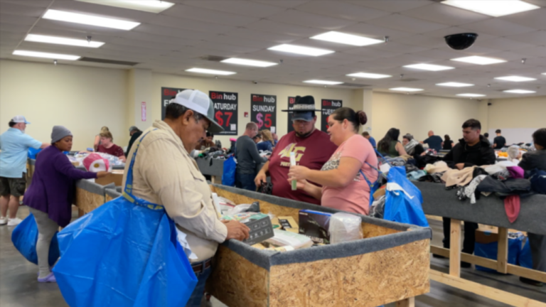 ChatGPT said: A group of people browsing through merchandise in large wooden bins at a busy bin store. Shoppers carry large blue shopping bags and examine various discounted items. Signs on the wall display daily pricing, such as "$7 Saturday" and "$5 Sunday," indicating a tiered pricing structure.