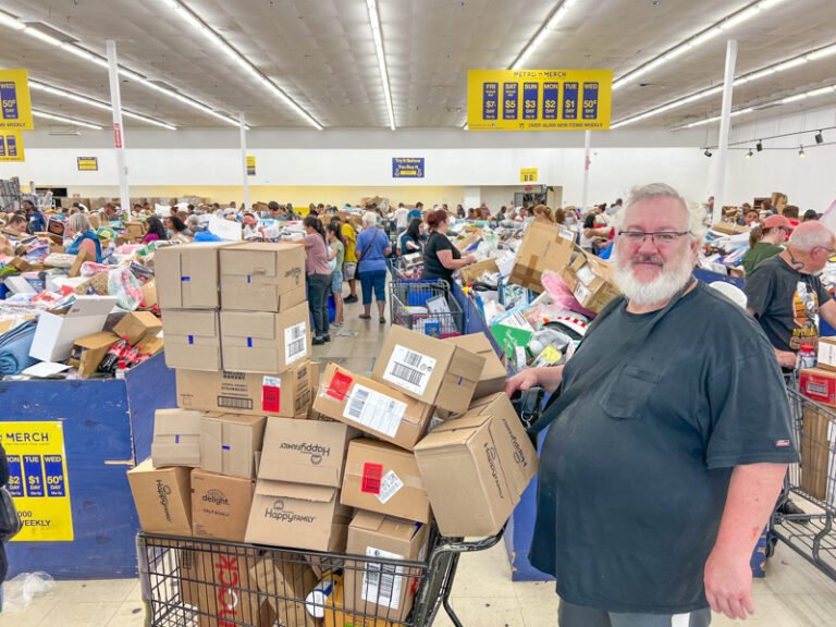 A bearded man in a black shirt stands next to a shopping cart stacked with boxes at a crowded bin store, where people are browsing through large blue bins filled with discounted merchandise.