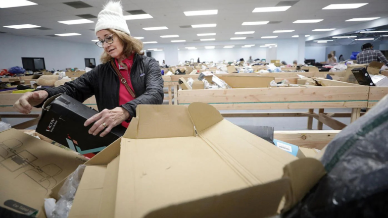 A woman wearing a white beanie and glasses examines a boxed item at a bin store filled with large sorting tables and discounted merchandise. The store is brightly lit, and various products are scattered across open bins in the background.