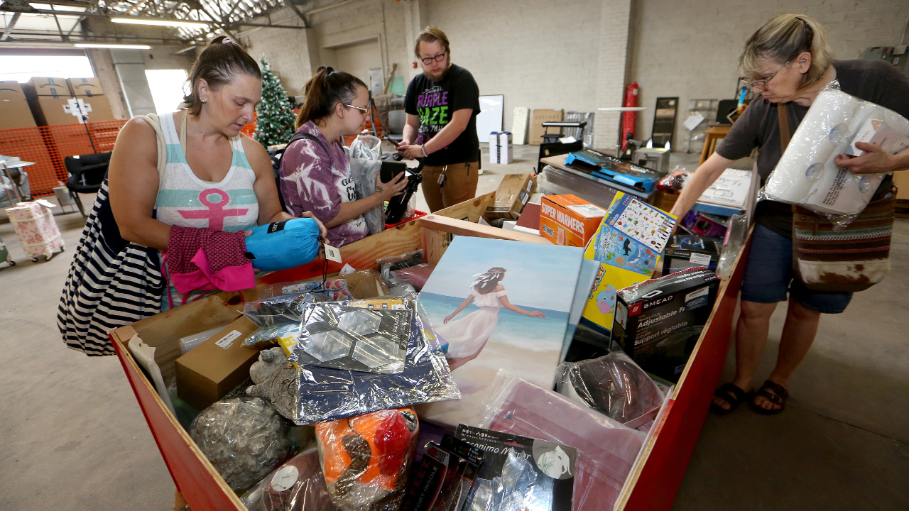 Shoppers browse through a bin filled with assorted discounted items, including electronics, artwork, clothing, and household goods at a busy bin store