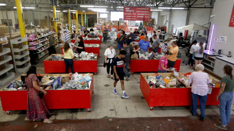 Shoppers browse through large red bins filled with assorted discounted merchandise inside a busy bin store with industrial-style lighting and shelving.