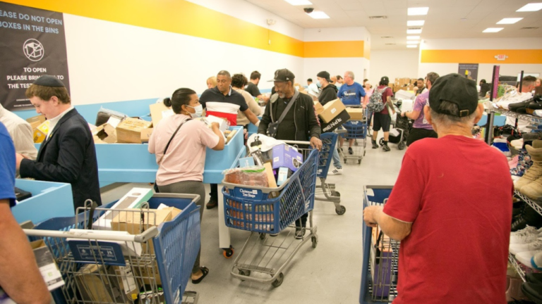 Shoppers browse through merchandise in blue bins and push carts filled with various items at a busy bin store, creating a lively and crowded retail environment.
