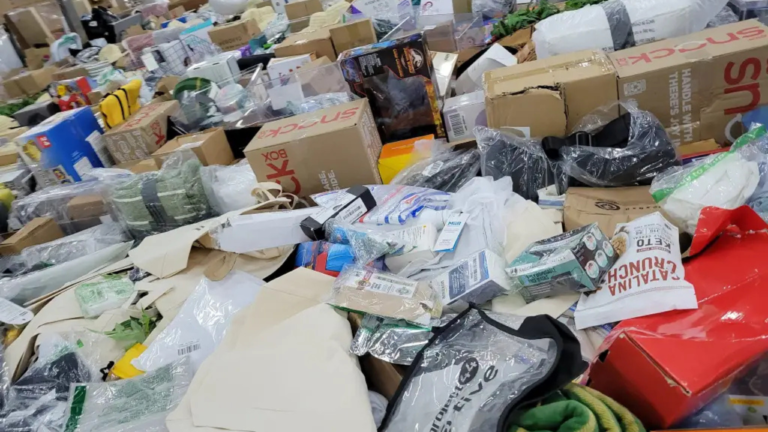 Close-up view of a bin filled with assorted products at a bin store, including boxed items, packaged snacks, clothing, and household goods.