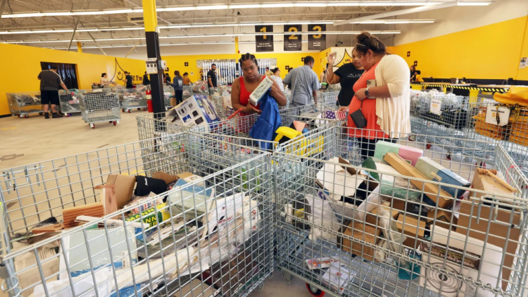 Shoppers browsing through large metal bins filled with assorted discounted merchandise inside a brightly lit bin store with yellow walls and signage.
